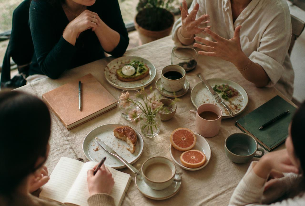 Overhead view of a brunch table with coffee, notebooks and hands mid-discussion